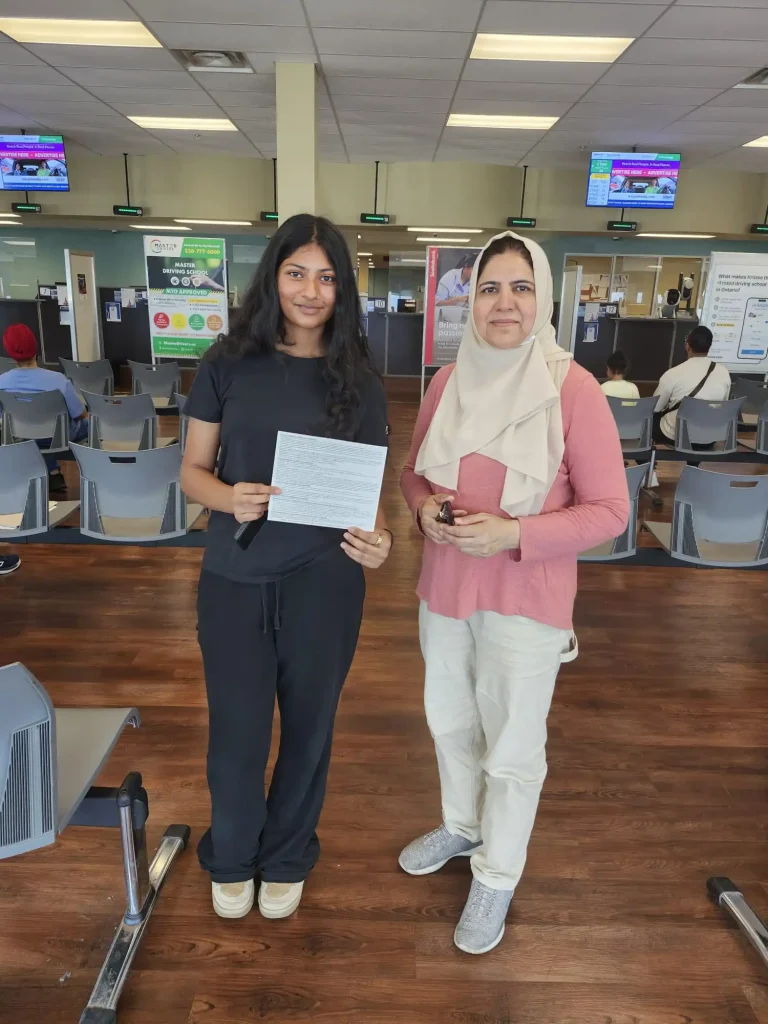 Student Emaan holding her passed G2 driver's license test paper next to her driving instructor Tayyaba Zahra inside a Mississauga DriveTest center.
